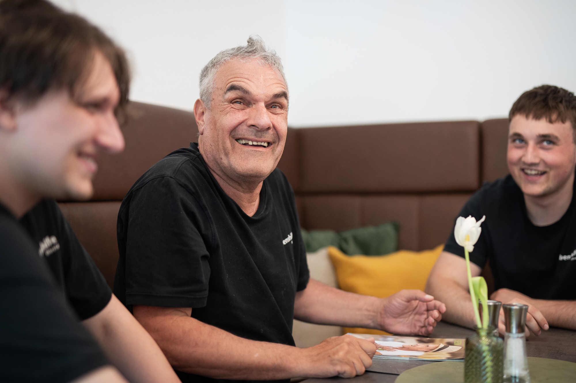 Ein Mann sitzt lachend auf einer Sitzbank am Tisch mit je links und rechts einem Mann, die ebenfalls lächeln