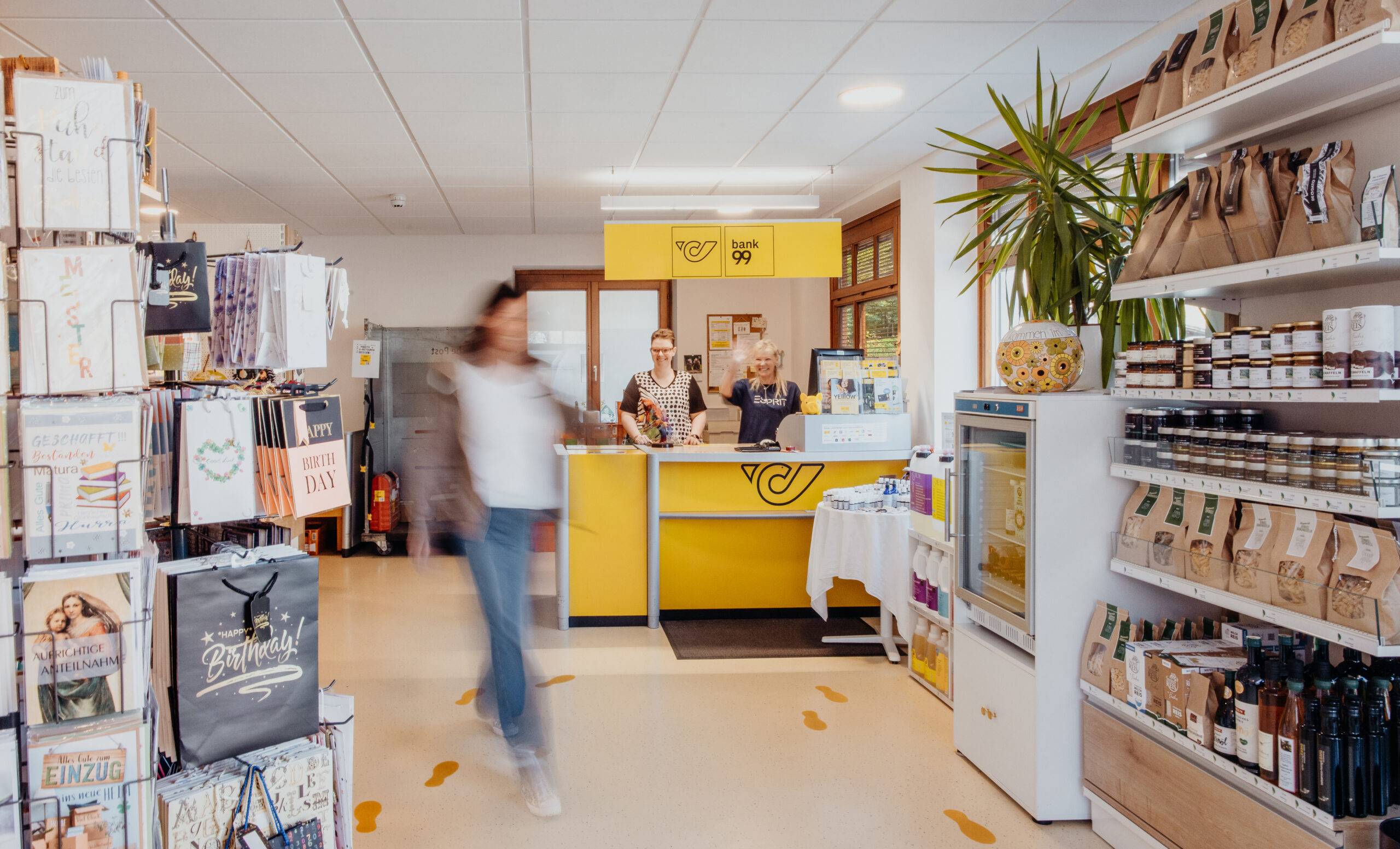 Zwei Frauen bedienen in der Post einen Postkunden und lächeln ihn an.