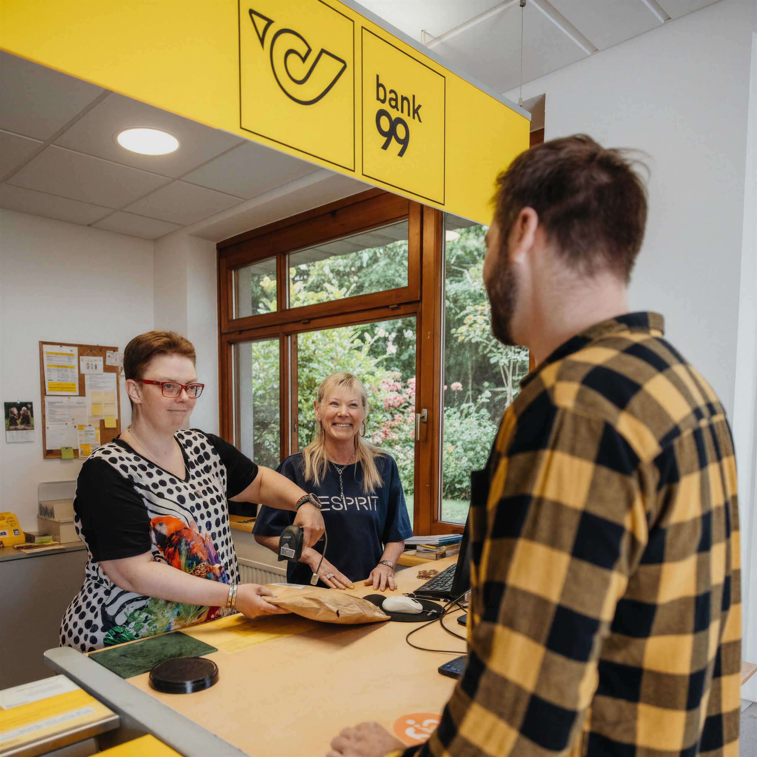 Zwei Frauen bedienen in der Post einen Postkunden und lächeln ihn an.