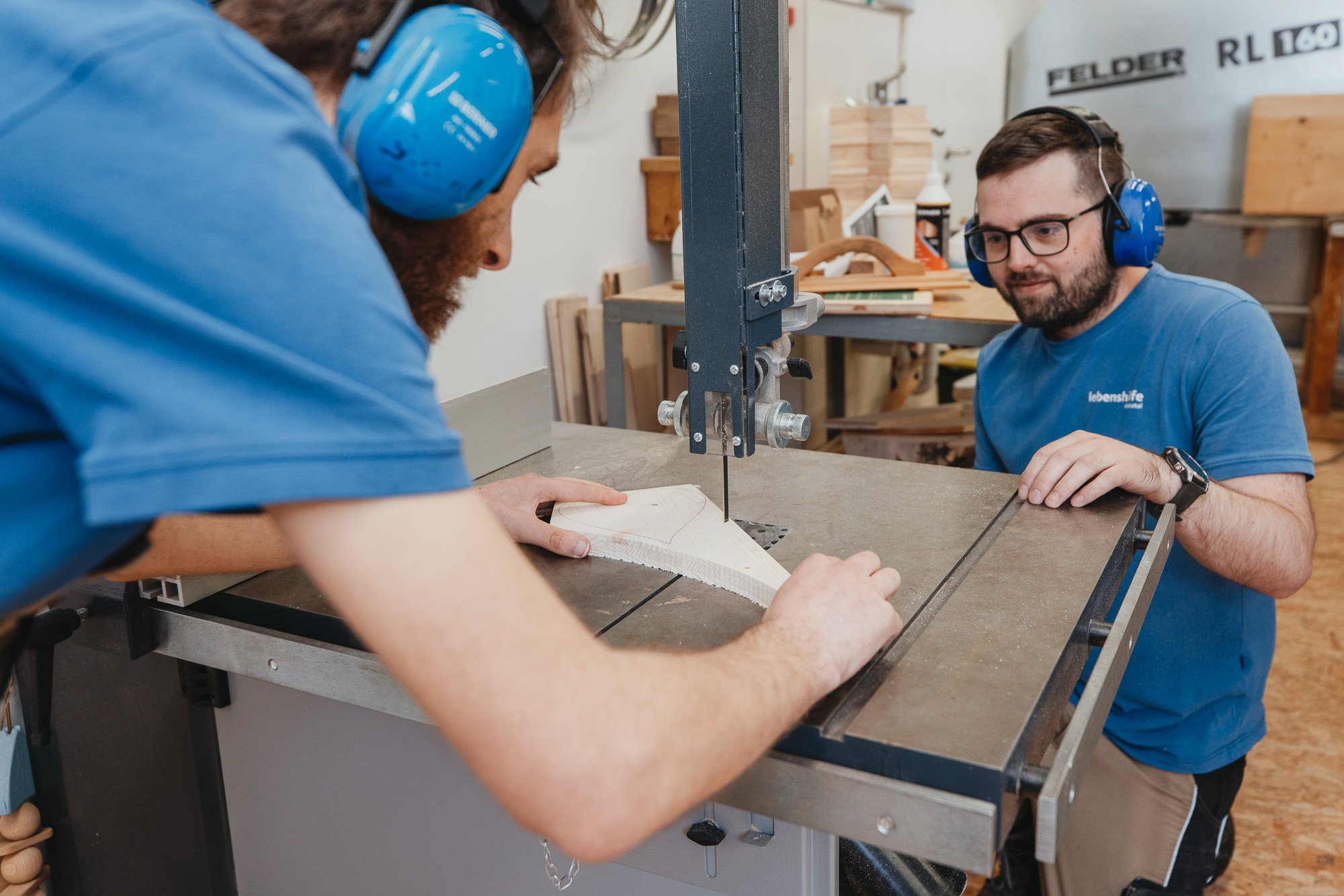 Zwei Männer arbeiten gemeinsam an einer Holzsäge und tragen Gehörschutz.