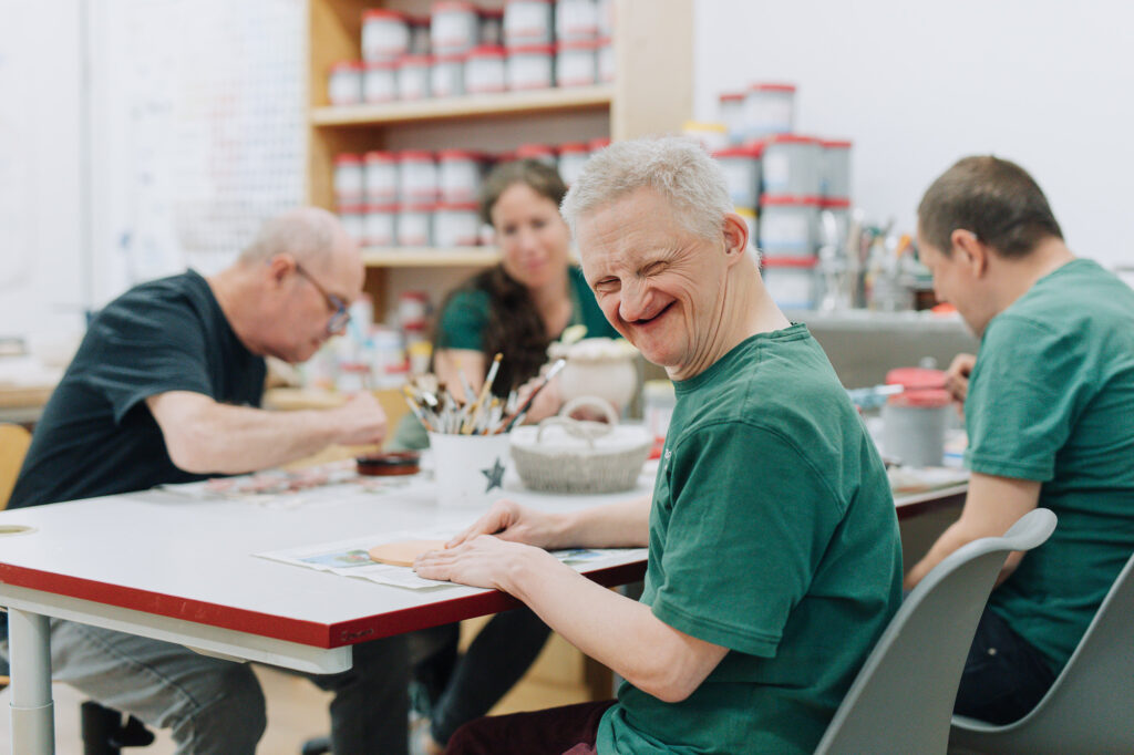 Ein in die Kamera lachender Mann sitzt an einem Tisch mit drei weiteren Personen und sie arbeiten gemeinsam an Bastelprojekten aus Ton.