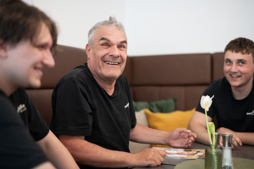 Ein Mann sitzt lachend auf einer Sitzbank am Tisch mit je links und rechts einem Mann, die ebenfalls lächeln