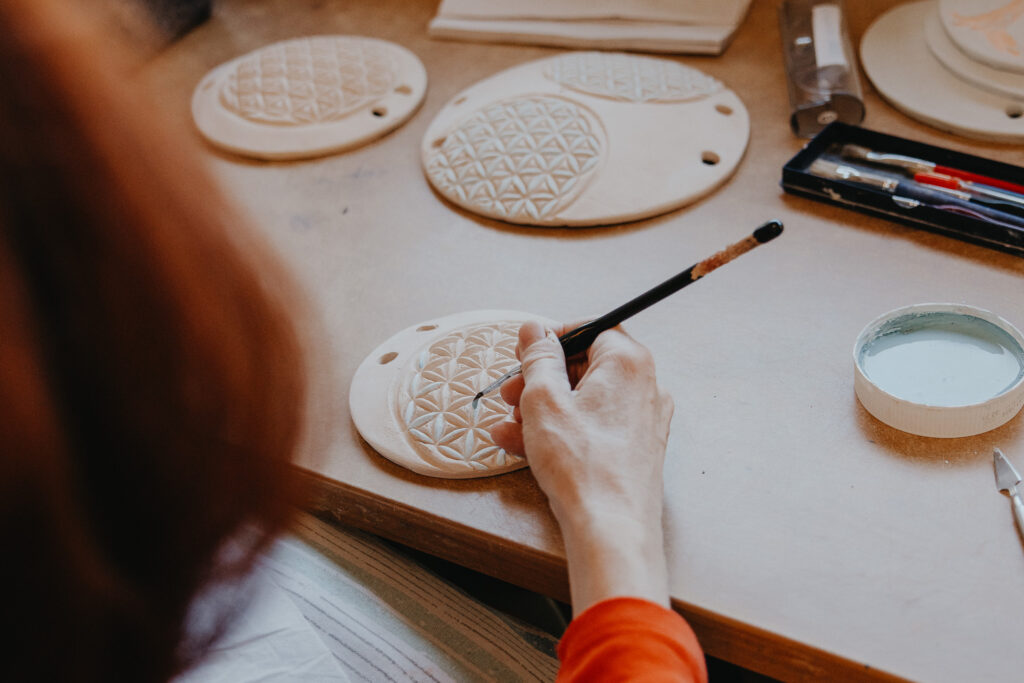 Eine Frau bemalt eine kunstvoll geformte Tonplatte mit einem Pinsel und graublauer Farbe.