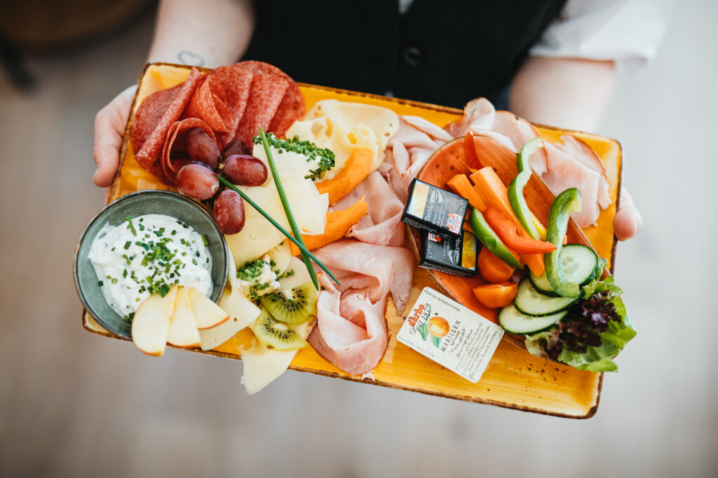 Jemand hält ein viereckiges, längliches, oranges Tablett, auf dem sich verschiedene Wurst- und Käsesorten, Gemüse, Obst und ein Aufstrich befinden.