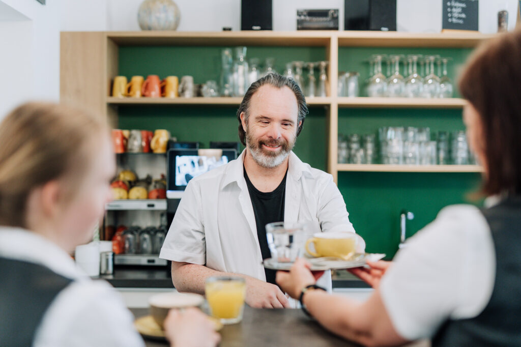 Ein Mann steht im Restaurant am Tresen und überreicht lächelnd ein Tablett mit Kaffee und einem Glas Wasser an eine Frau als Bedienung.