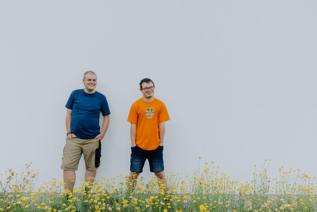 Zwei Männer stehen lächelnd an der Hauswand und haben die Hände eingesteckt. Davor ist eine Blumenwiese.