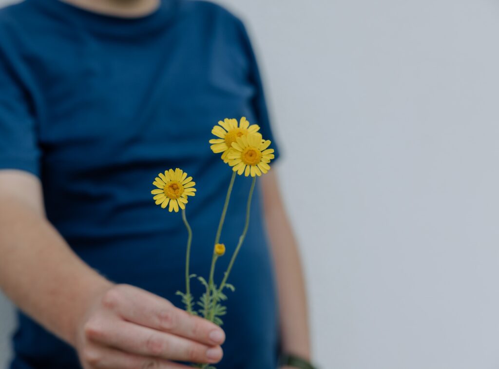 Lebenshilfe Muerztal_WEB (C) nicoleseiser-108 - gedreht Ein junger Mann mit blauem T-Shirt streckt die linke Hand aus. Er hält eine Blume mit drei gelben, leuchtenden Blüten in der Hand.